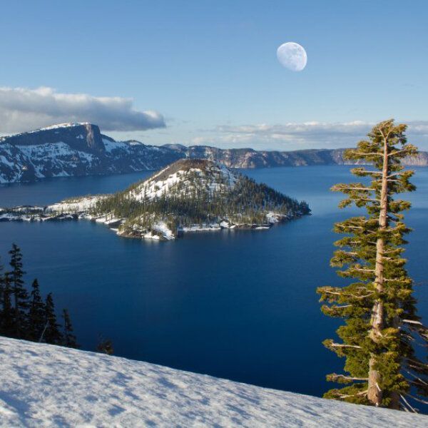 Vue sur le lac Crater enneigé avec la lune dans le ciel bleu.
