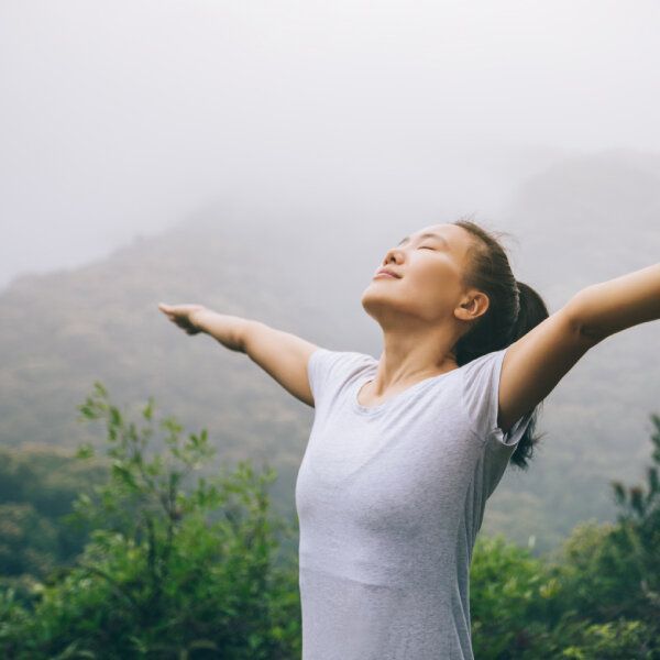 Une jeune femme écarte les bras et prends une grande inspiration devant un arrière-plan de forêt brumeuse.