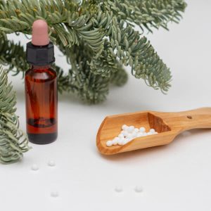 Devant des feuilles de pin, un flacon et une cuillère en bois remplie de granulés homéopathiques.