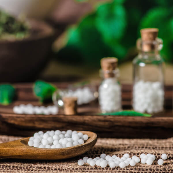 Une cuillère en bois remplie de granules homéopathique avec des flacons de granules en arrière-plan.